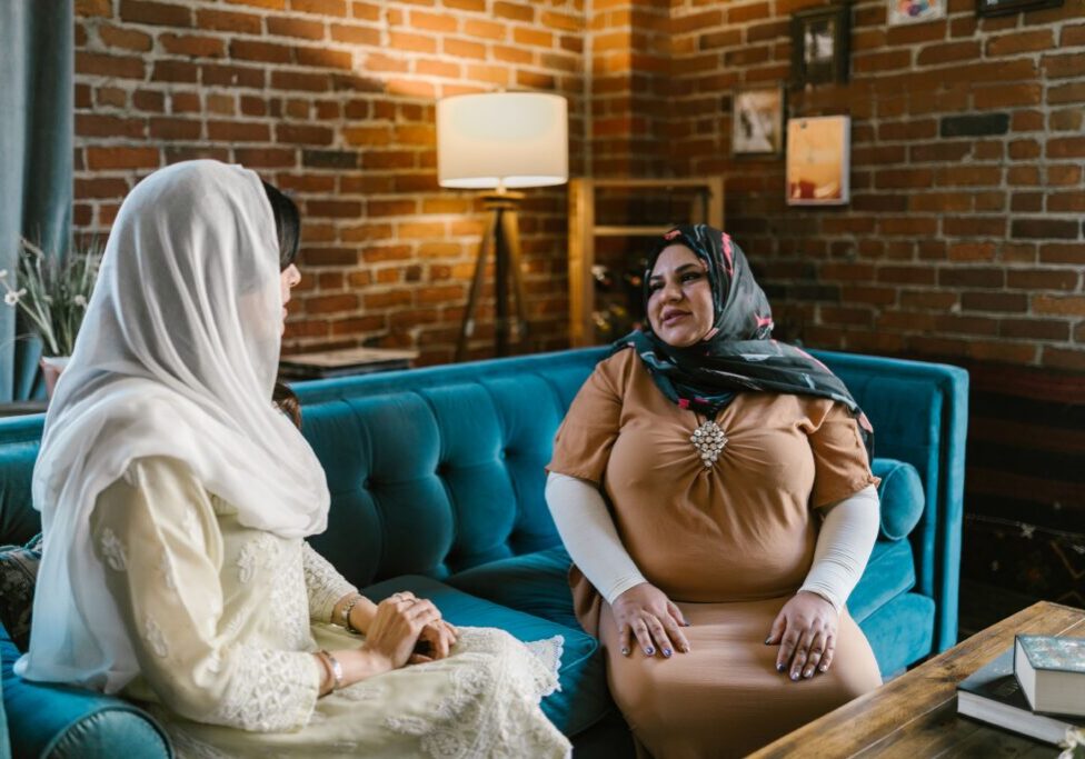 Two women in hijabs sharing a warm conversation in a cozy, stylish interior with brick walls.